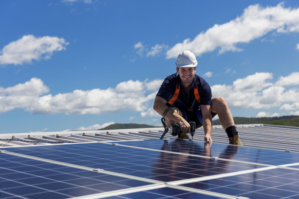 Solar panel installer with drill installing solar panels on a commercial roof on a sunny day.